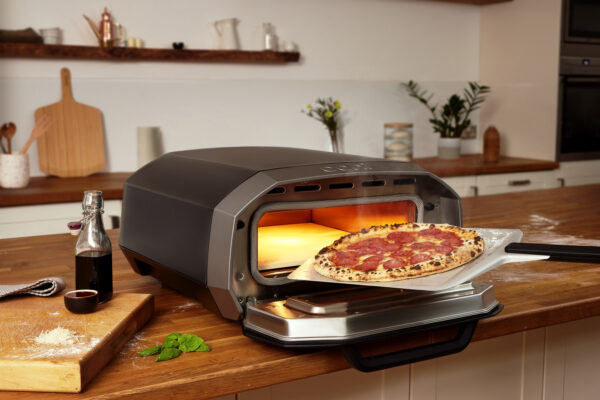 Ooni Volt 12 Electric Pizza Oven - Inside on wooden kitchen bench. A Neapolitan Pizza is coming out of the Ooni Volt 12 fully cooked after just 90 seconds, we can see that the oven is glowing red hot at 450 degrees Celsius.