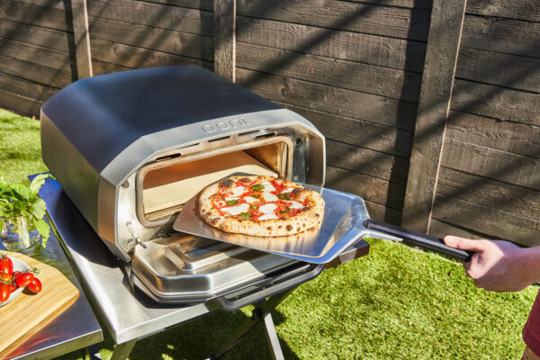 Ooni Volt 12 Electric Pizza Oven - Outside on a table on a lawn, fence in background. A Neapolitan Pizza is coming out of the Ooni Volt 12 fully cooked after just 90 seconds.